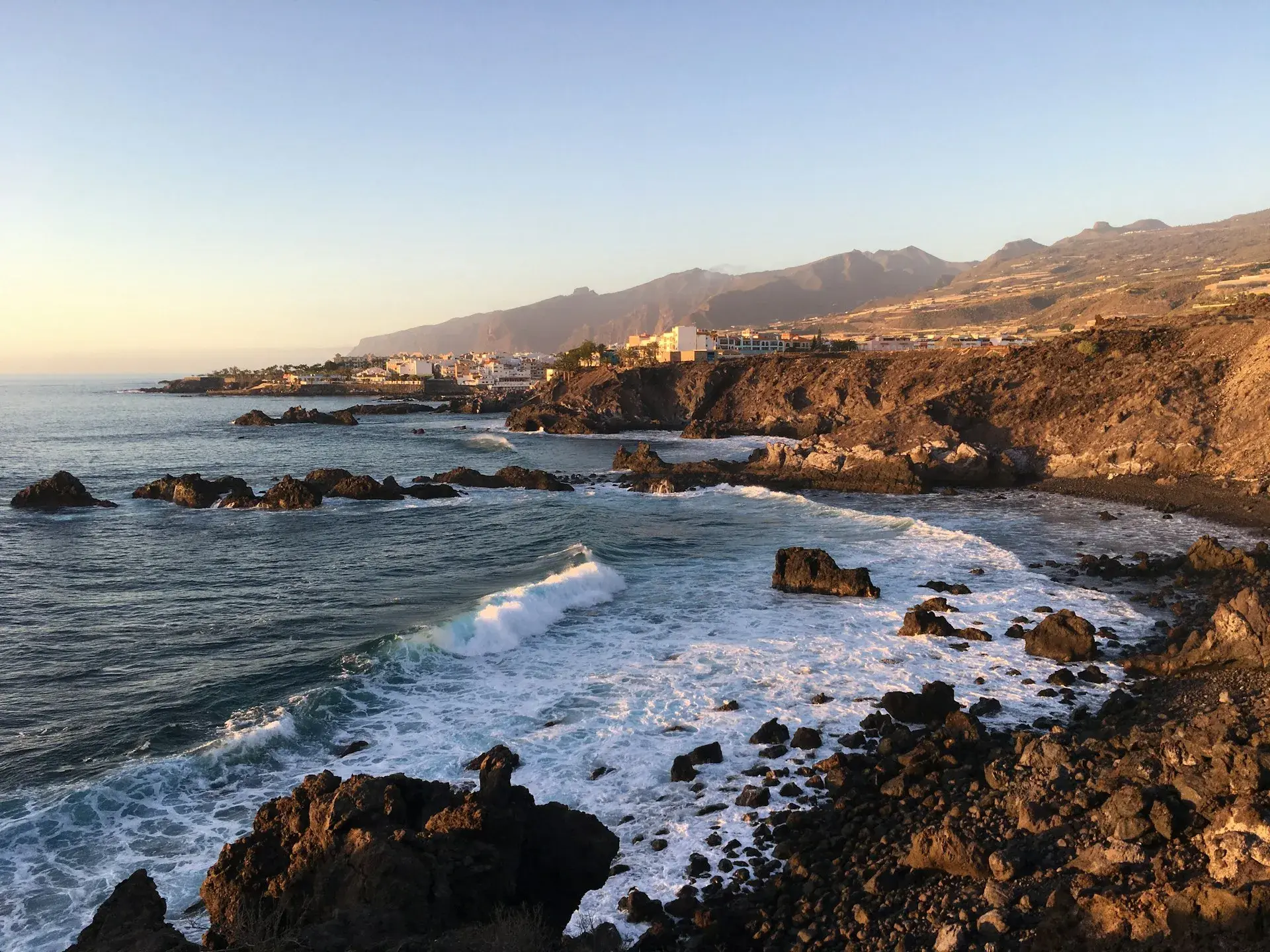 Costa rocosa del norte de Tenerife con olas rompiendo y un pueblo junto al mar al atardecer