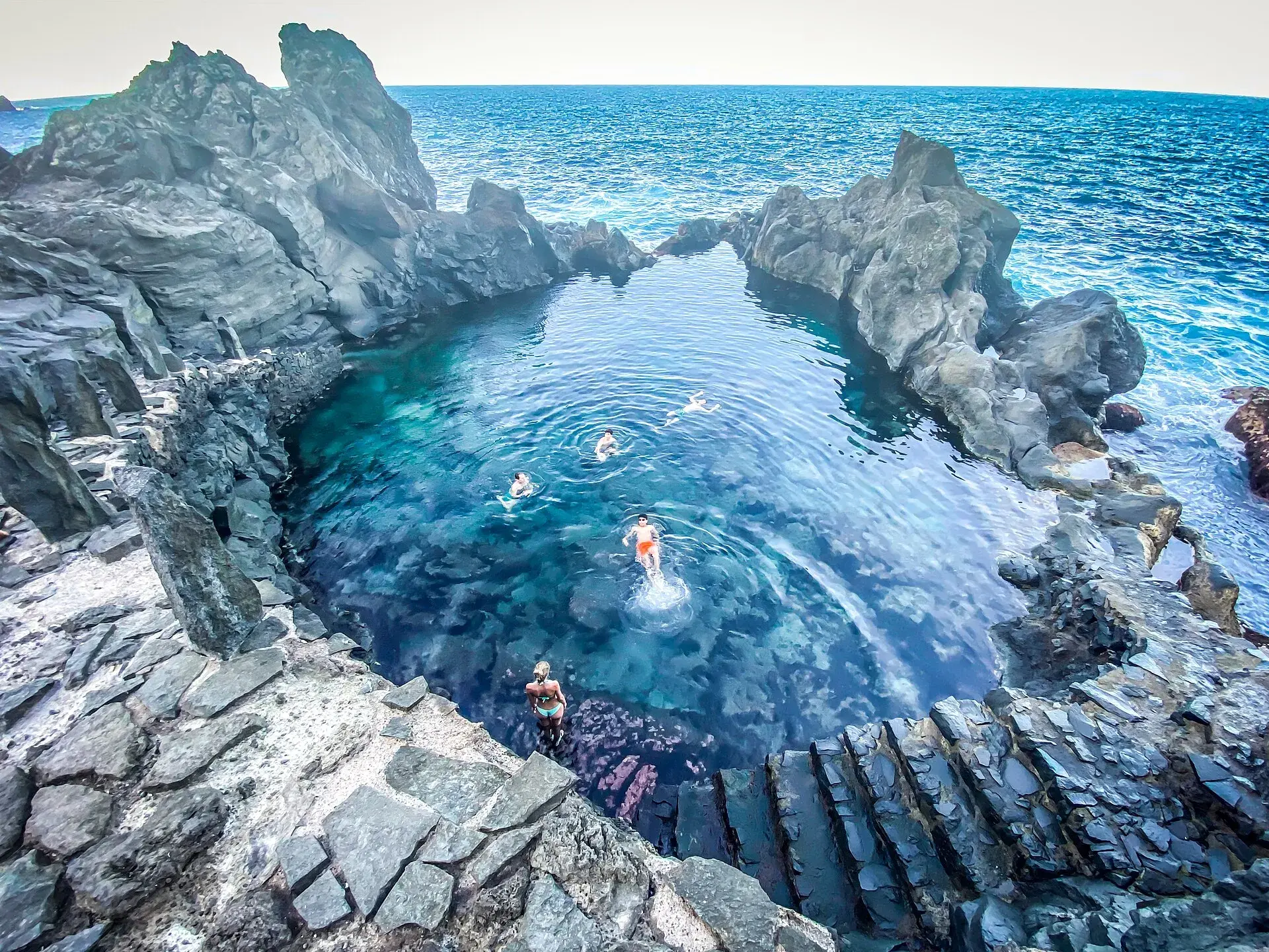 Charco natural de agua turquesa entre rocas volcánicas en la costa de La Guancha, con bañistas y el océano al fondo