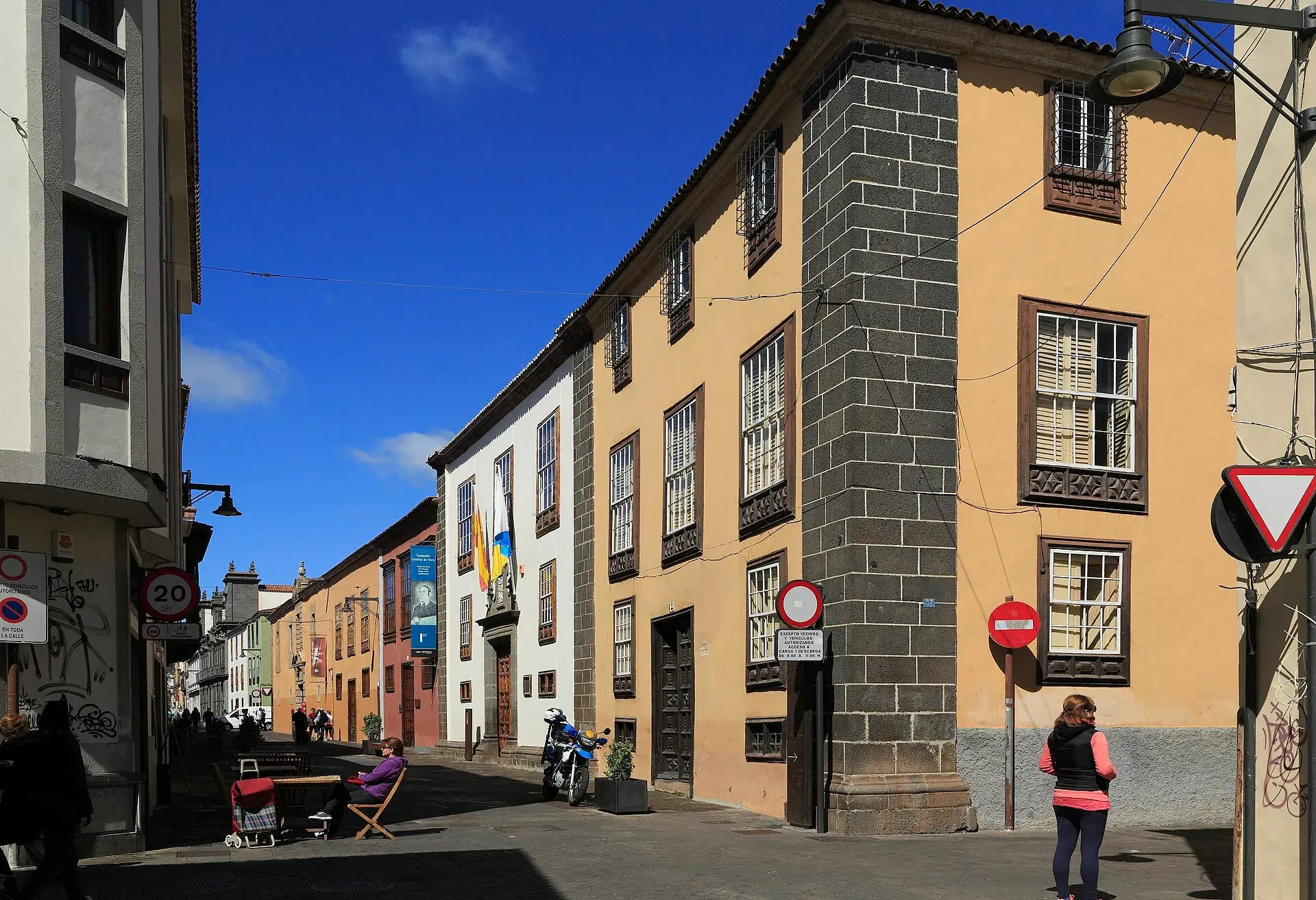 Calle San Agustín in the historic centre of San Cristóbal de La Laguna, with Canarian mansions in ochre and white, wooden windows, a dark stone corner and pedestrians under a blue sky