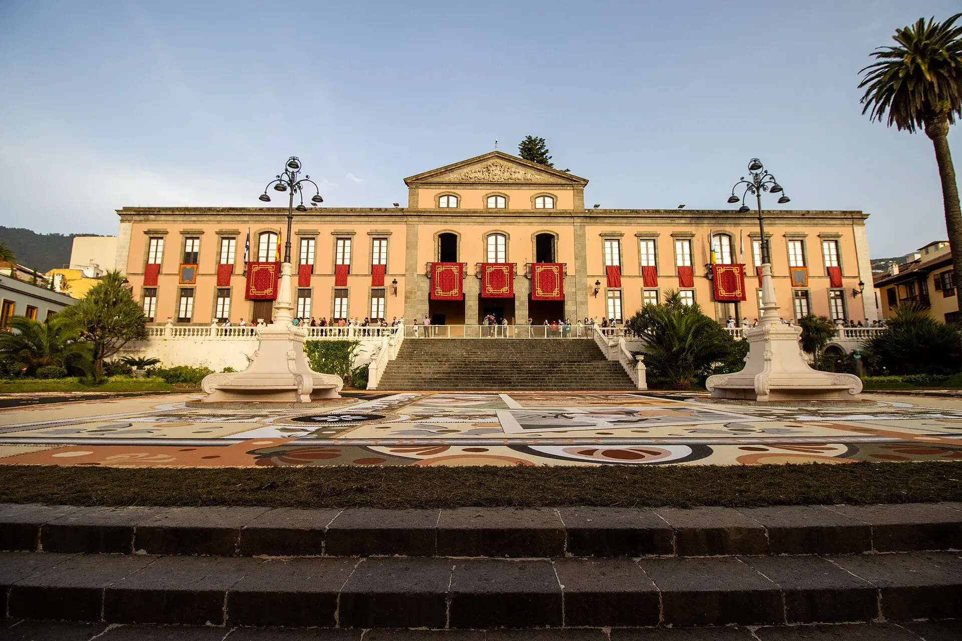 Fachada neoclásica del Ayuntamiento de La Orotava con colgaduras rojas en los balcones y, en la plaza, la gran alfombra de arena volcánica de colores del Corpus, vista desde unas escaleras de piedra
