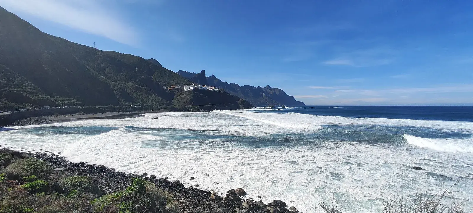 Oleaje sobre la playa de Almáciga, con un caserío en el promontorio y los picos de Anaga al fondo