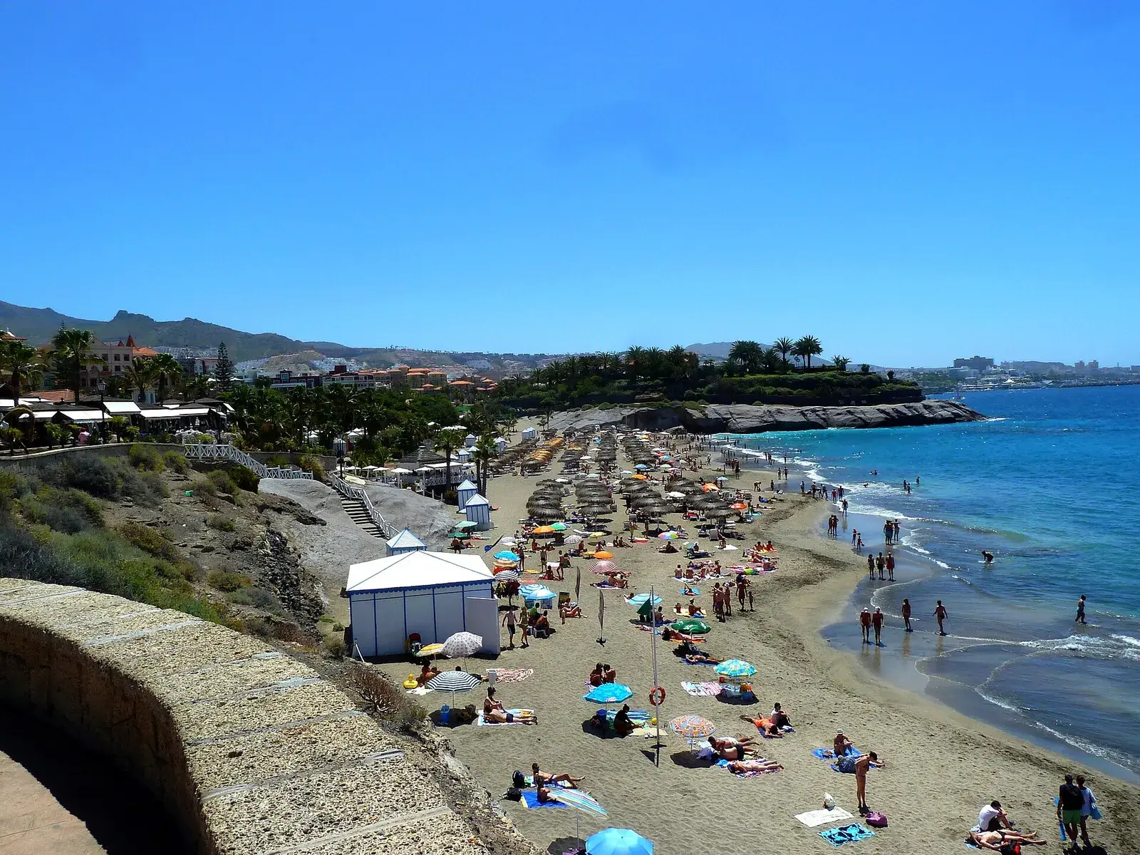 Playa del Duque desde el paseo, con sombrillas de paja, casetas blancas y el mar turquesa