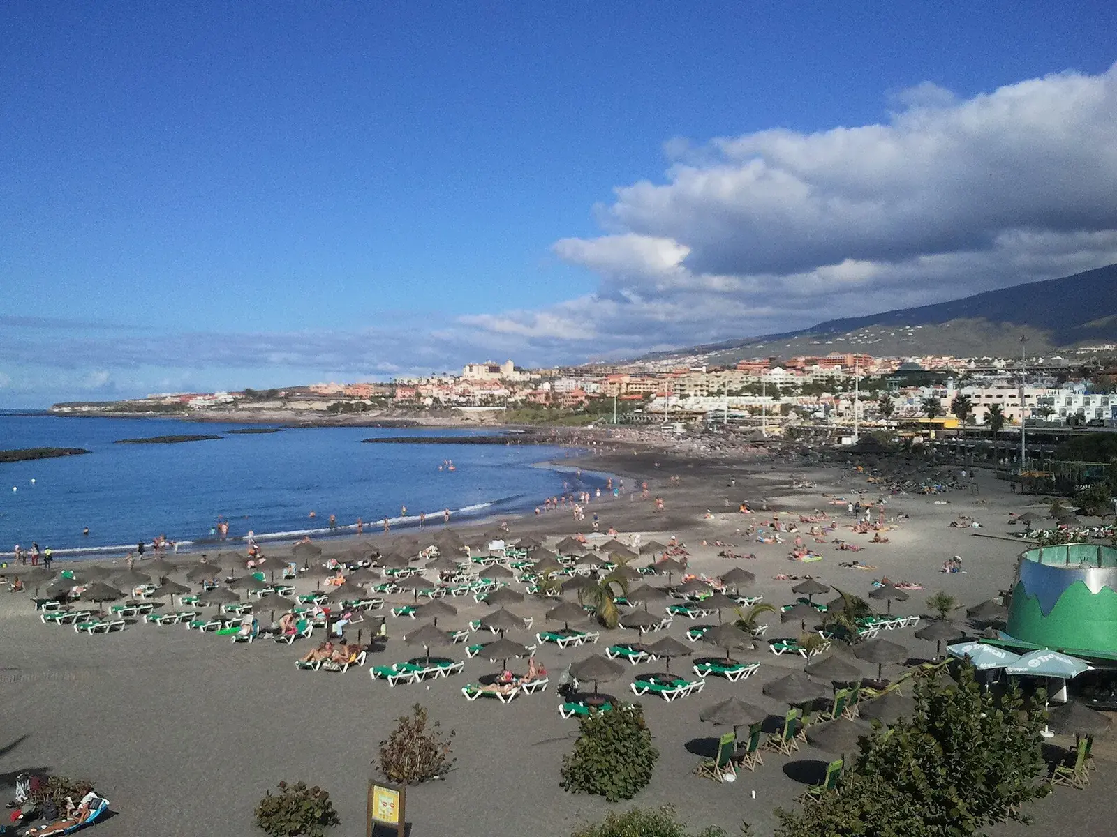 Playa de Fañabé desde el paseo, con sombrillas de paja, hamacas verdes y Costa Adeje al fondo