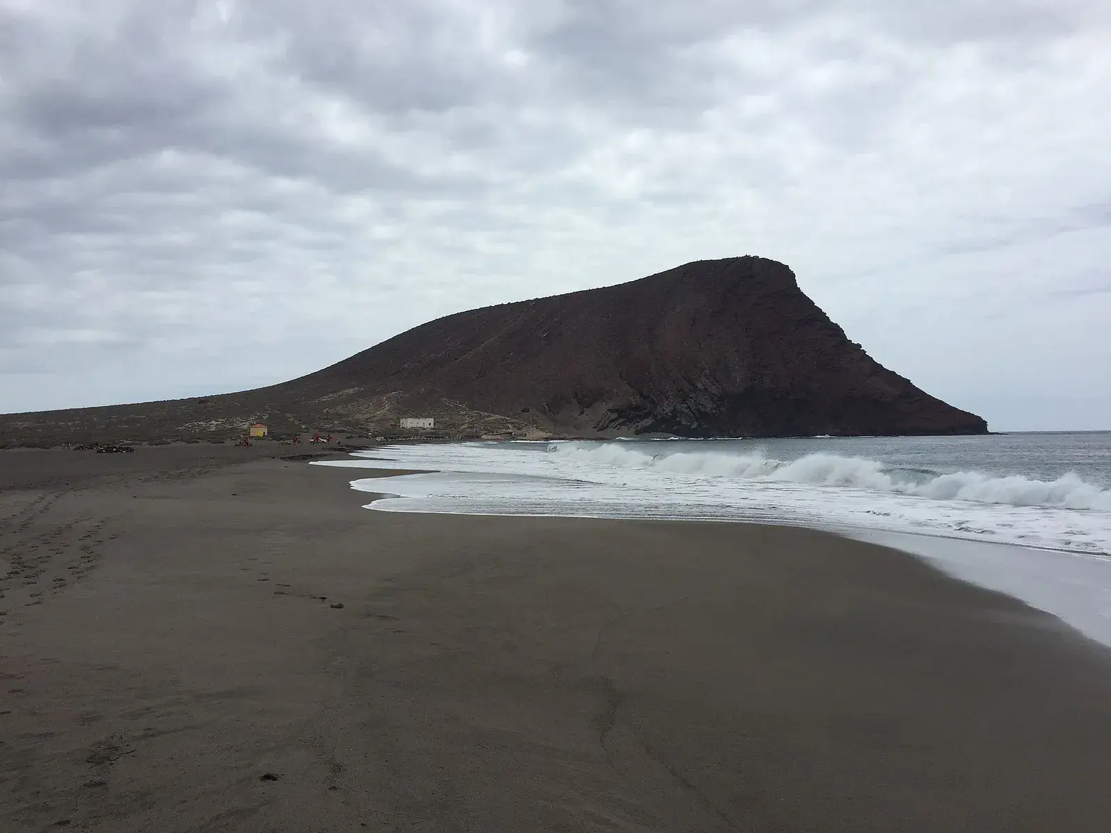 Playa de La Tejita con la arena mojada, olas en la orilla y la Montaña Roja bajo un cielo nublado