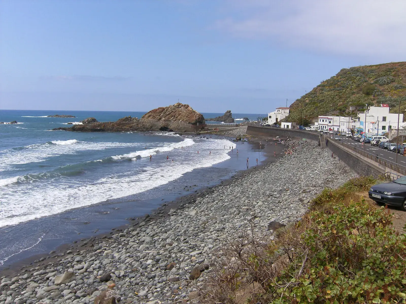 Playa de arena oscura y callaos de Roque de las Bodegas, con el roque en el mar y casas blancas junto a la carretera