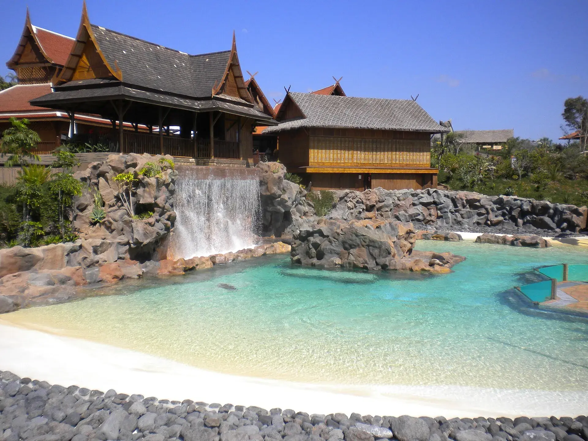 Thai-style wooden pavilions and a waterfall above a turquoise pool with white sand at Siam Park, Costa Adeje