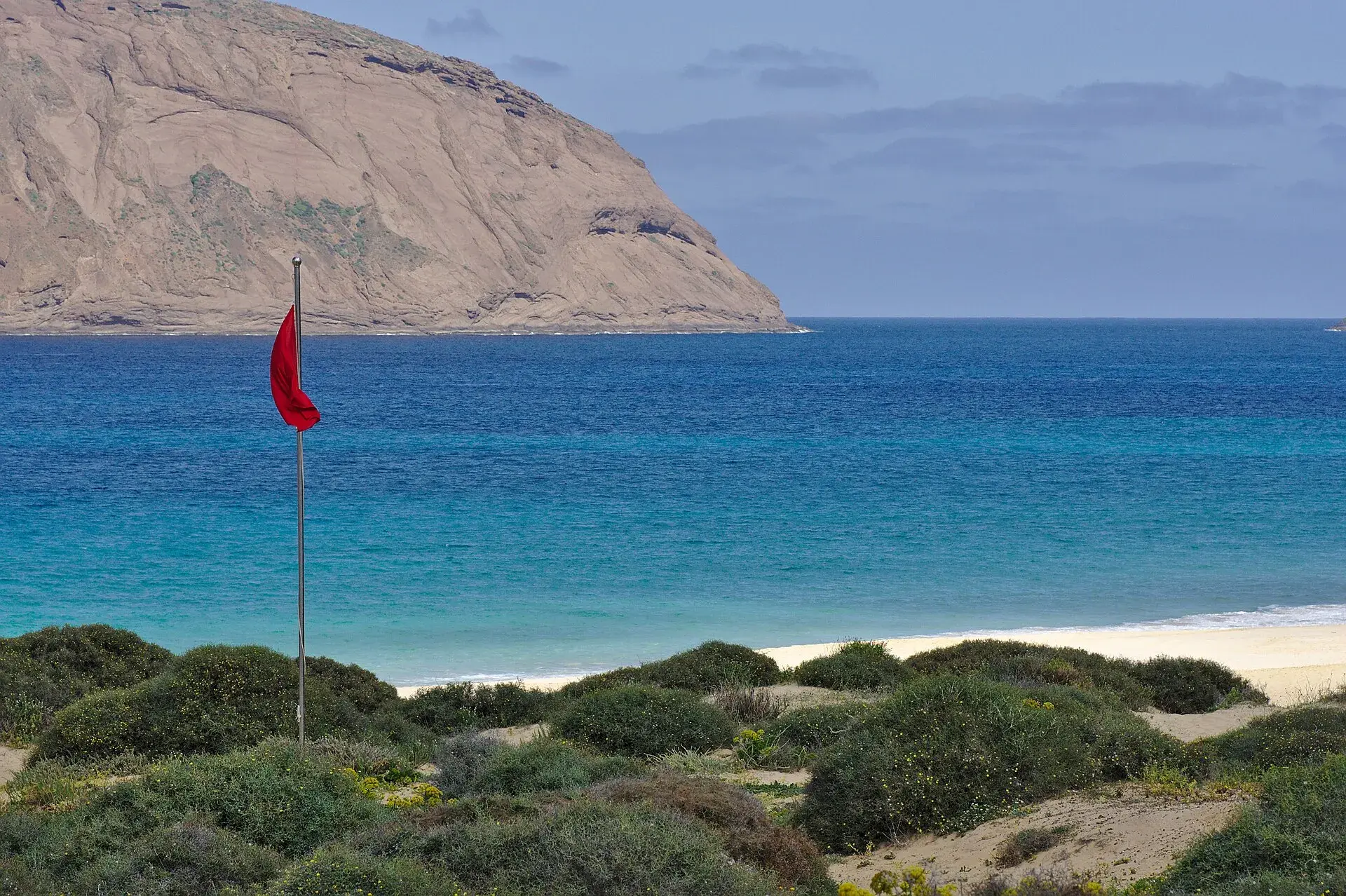Bandera roja ondeando junto a una playa de arena dorada de Canarias, con el mar turquesa y un risco al fondo